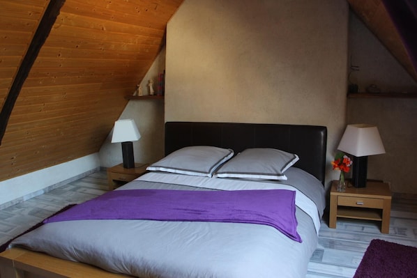 Un dormitorio con techo inclinado, una cama con manta morada, dos mesitas de noche con lámparas y un pequeño florero con flores.