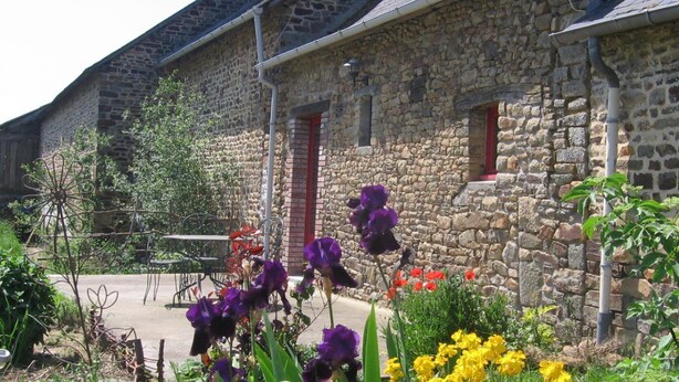 A stone building with a red door and windows, surrounded by a garden with purple and yellow flowers. - Aux Bonnes Herbes (Bais)