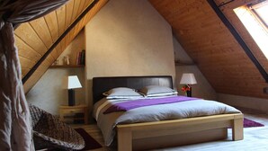 A bedroom with a sloped ceiling, a bed with a headboard, bedside lamps, a small table, and a chair. - Aux Bonnes Herbes (Bais)