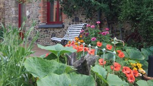 A stone building with a red-framed window, a wooden bench, and a garden with various flowers. - Aux Bonnes Herbes (Bais)
