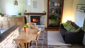 A cozy living room with a fireplace, a dining area with a wooden table and chairs, and a bookshelf filled with books and plants. - Aux Bonnes Herbes (Bais)