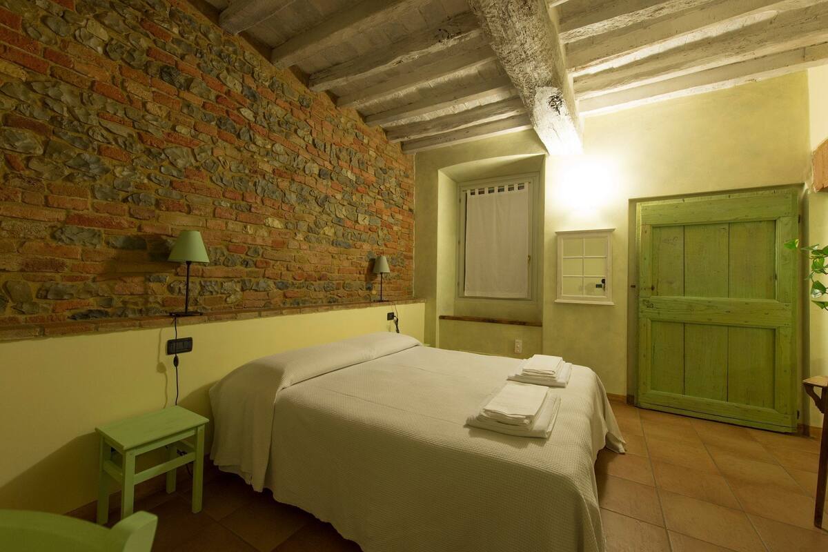 A bedroom with a brick wall, a bed with white linens, a green door, and a small table with a lamp.