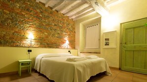 Ein Zimmer mit einem Bett, einem kleinen Tisch, einer Ziegelwand, einer Holztür und einer Lampe.
