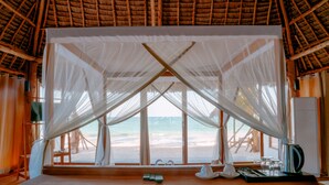 Vista a la playa desde una habitación con una mesa de madera, una cama y techo de paja.