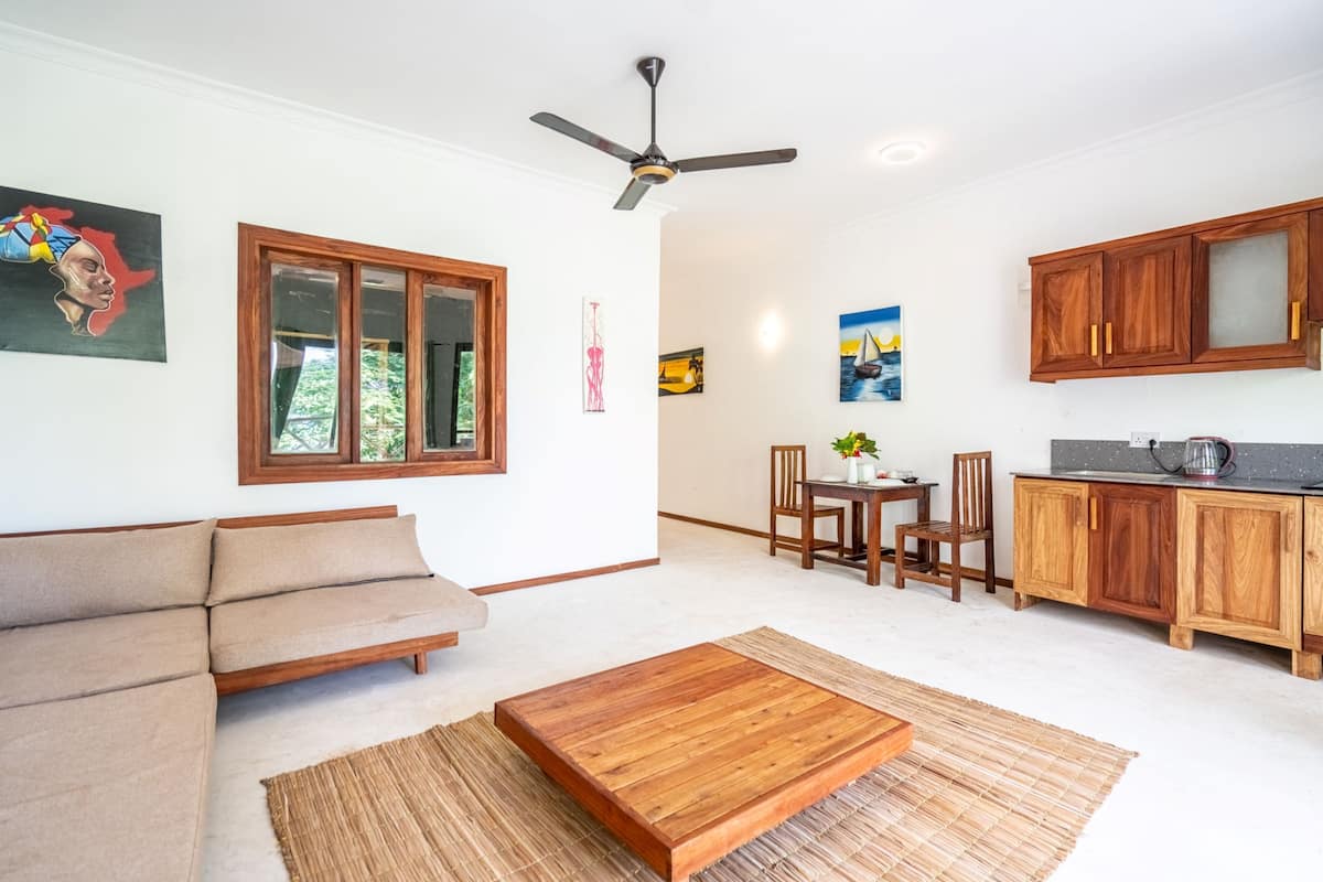 Una sala de estar moderna con un sofá, una mesa de centro de madera, y un comedor con sillas y una mesa. Hay una cocina con armarios de madera y un ventilador de techo.