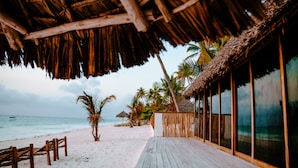 Una terraza de madera que conduce a una playa con una estructura de techo de paja, palmeras y vistas al océano.