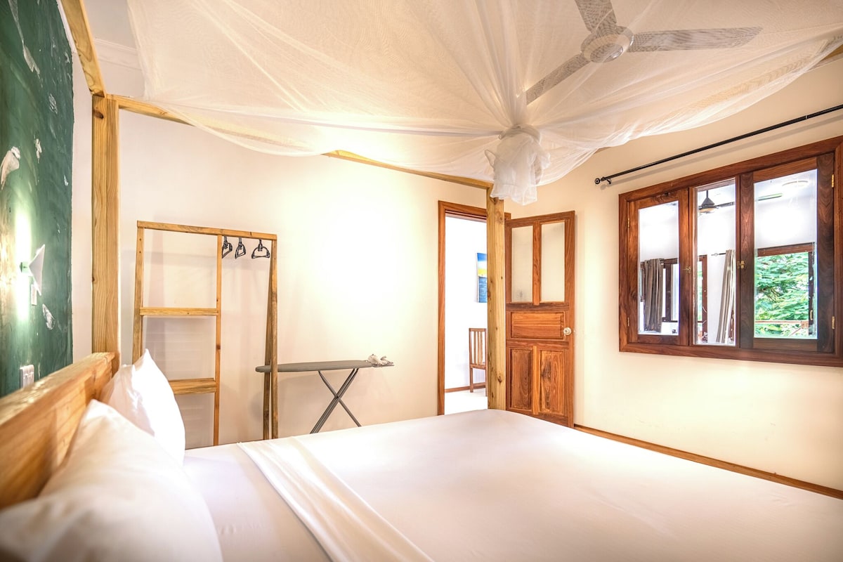 A room with a bed, a wooden wardrobe, a drying rack, and a window with a view of greenery.
