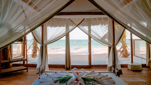 Vista a la playa desde una habitación con toldo, una cama, un banco y una pequeña mesa.