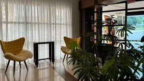 Eine Hotelhalle mit zwei gelben Sesseln, einem schwarzen Tisch und einem großen Fenster mit durchscheinenden Vorhängen.