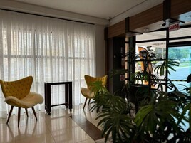 Um hall de hotel com duas cadeiras amarelas, uma mesa preta e uma janela grande com cortinas leves.