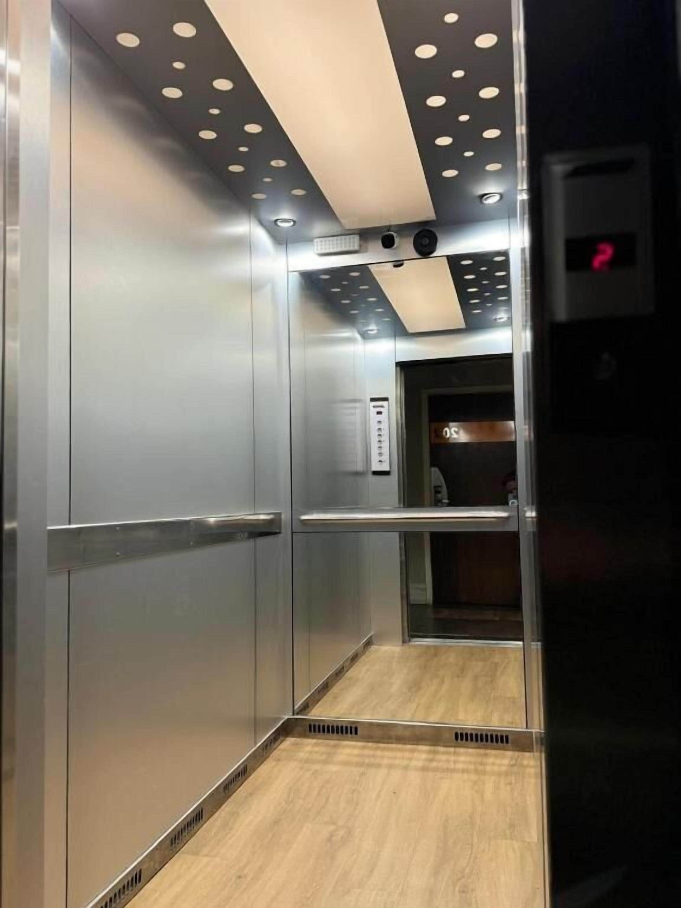 Interior de un ascensor con paredes metálicas, piso de madera y techo con lámparas circulares.
