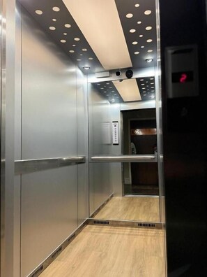 An elevator interior with metallic walls, a wooden floor, and a ceiling with circular light fixtures.