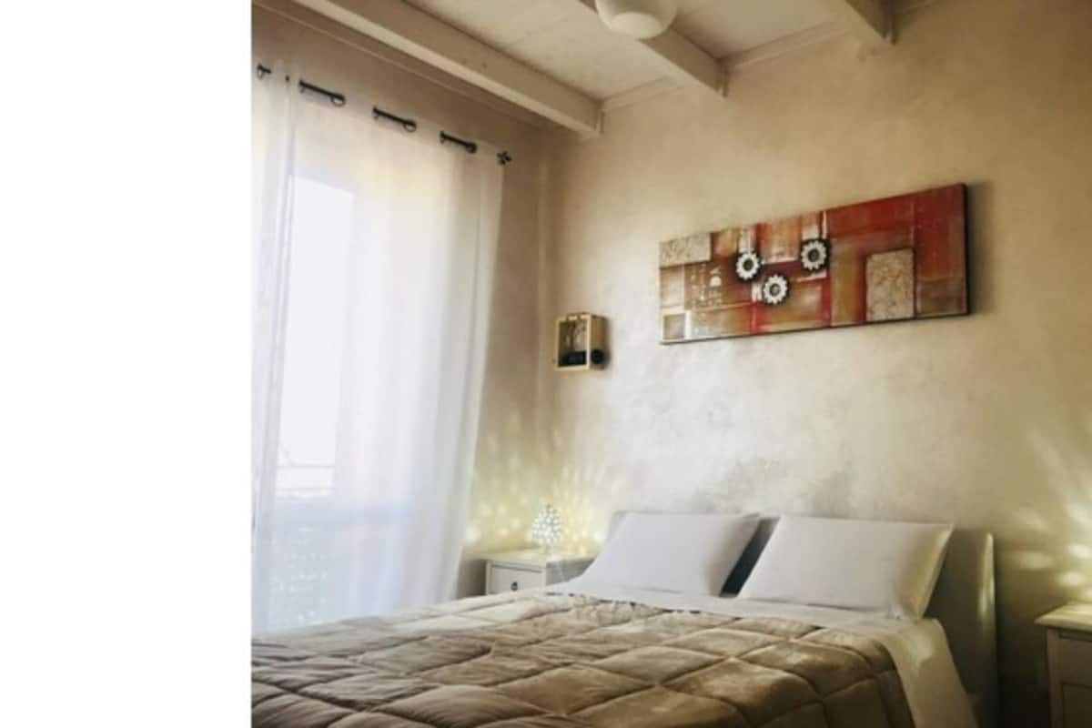 Ein Schlafzimmer mit einem Bett, einem Nachttisch, einer Wandleuchte und einem Fenster mit Vorhängen.