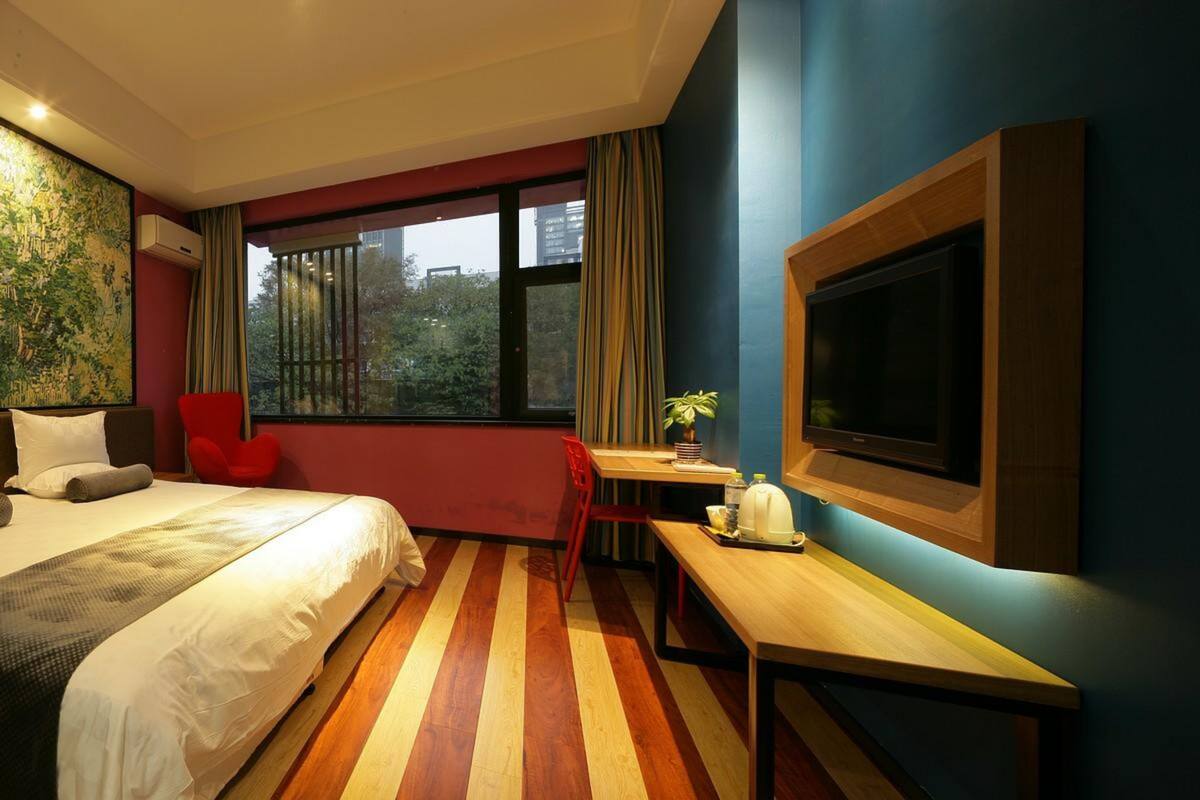 Une chambre d’hôtel équipée d’un lit, d’un bureau, d’une chaise, d’une télévision et d’une fenêtre donnant sur des immeubles.