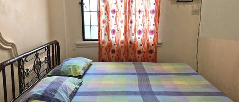 A single bed with a colorful checkered bedspread and two pillows, a window with a grid pattern, and an air conditioning unit on the wall.