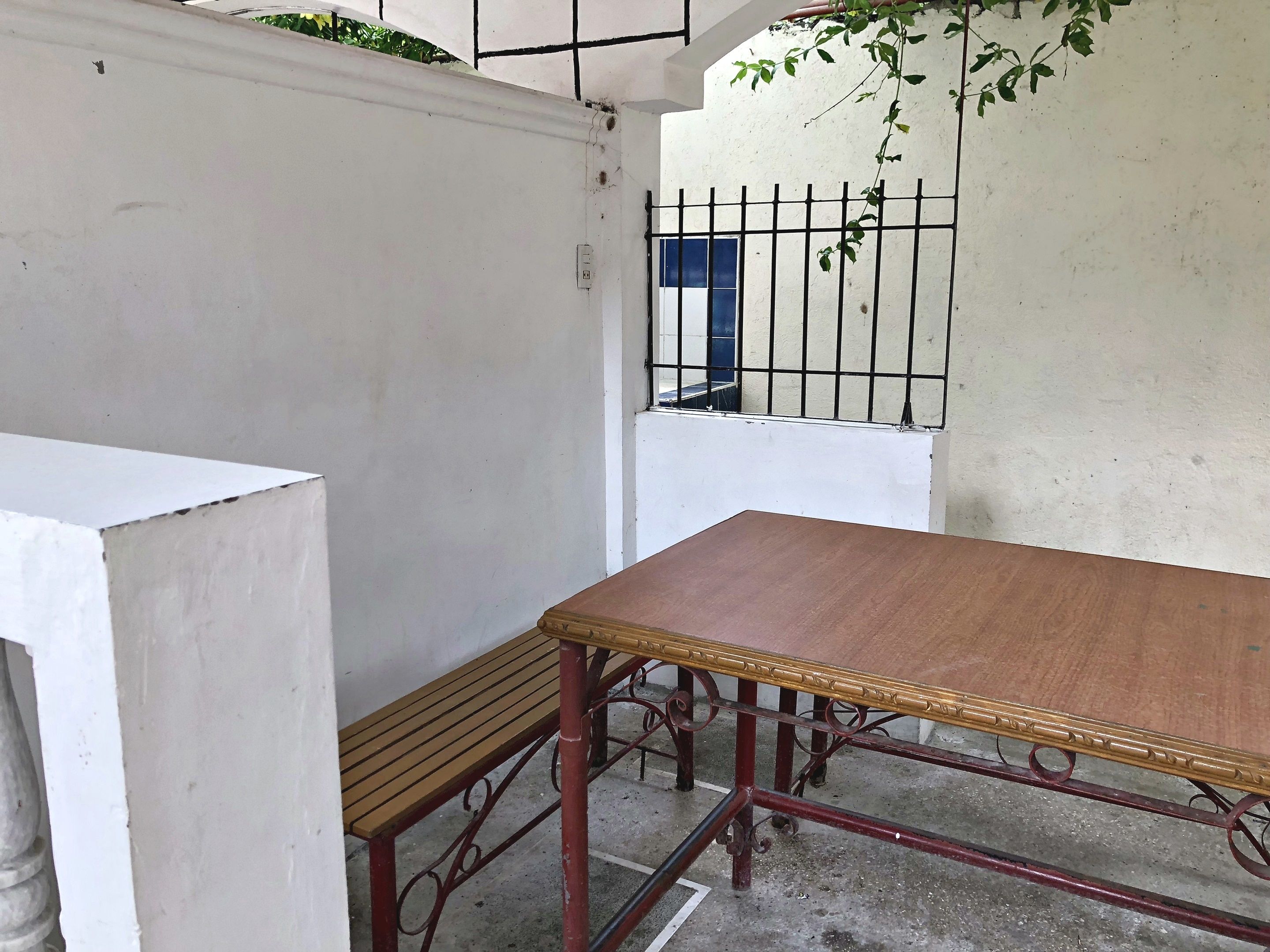 A corner of a room with a wooden table and bench, a white wall, a metal gate, and a small window.