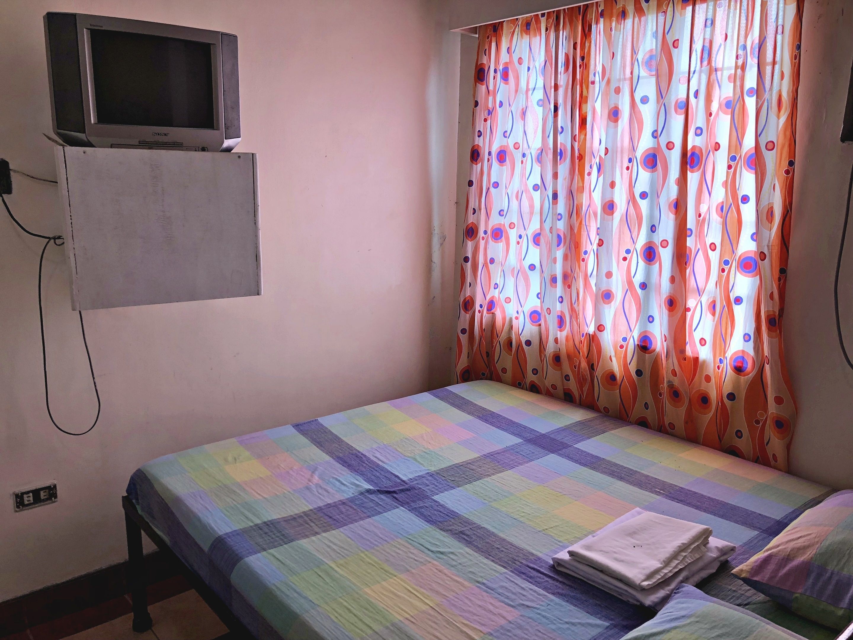 Una habitación con una cama, un televisor montado en un mueble y un ventanal con cortinas coloridas.