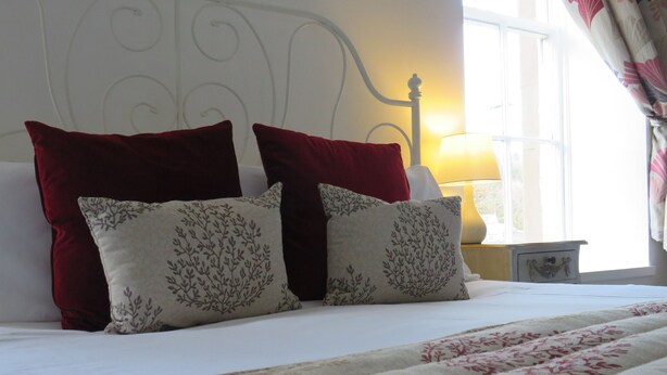 Ein ordentlich bezogenes Bett mit einer floralen Steppdecke und zwei Kissen, eines rot und eines mit einem Baum-Motiv, in einem Zimmer mit einem weißen Kopfteil und einer Nachttischlampe.