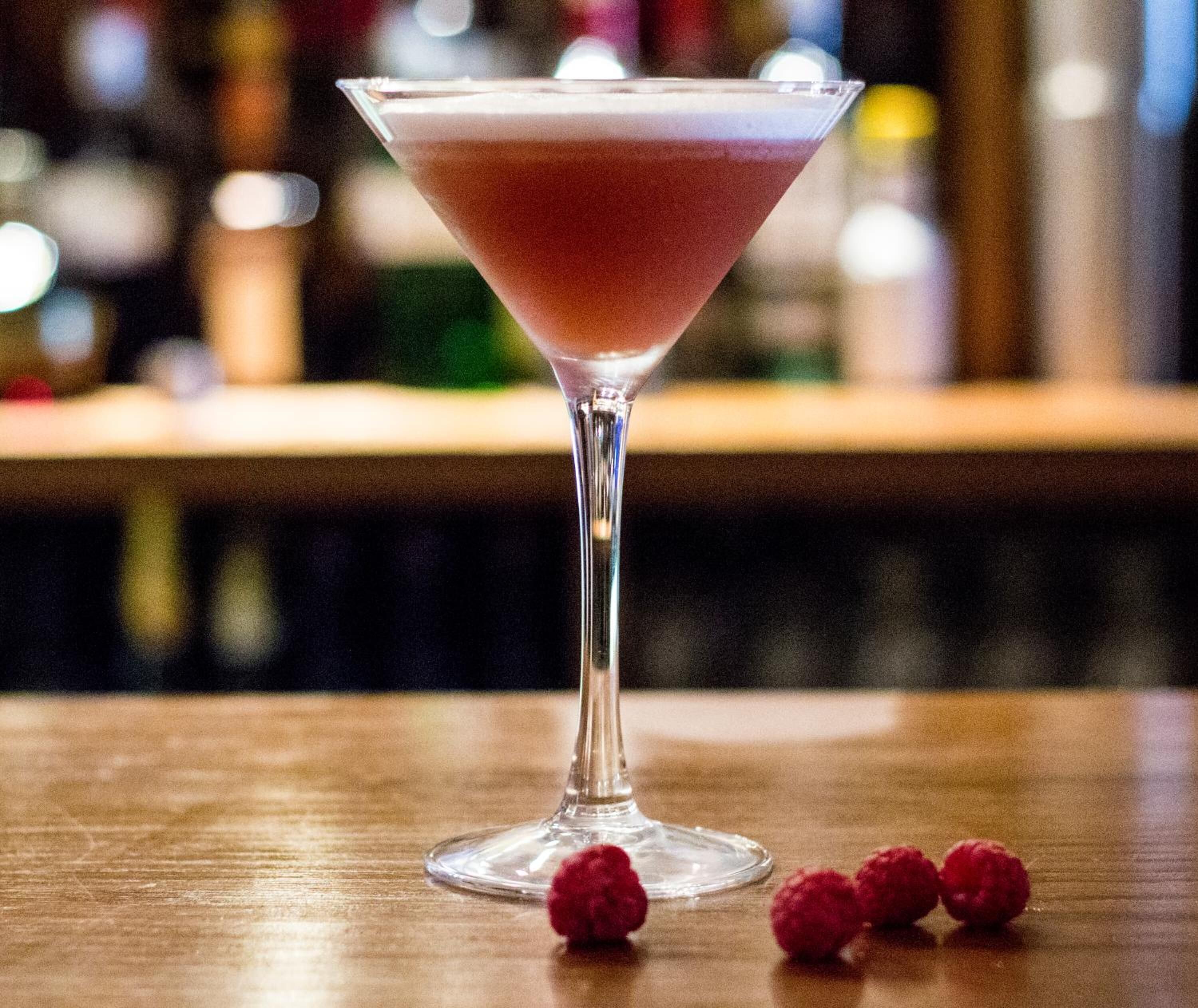 Una copa de martini con una bebida rosada sobre una barra de madera, acompañada de frambuesas.