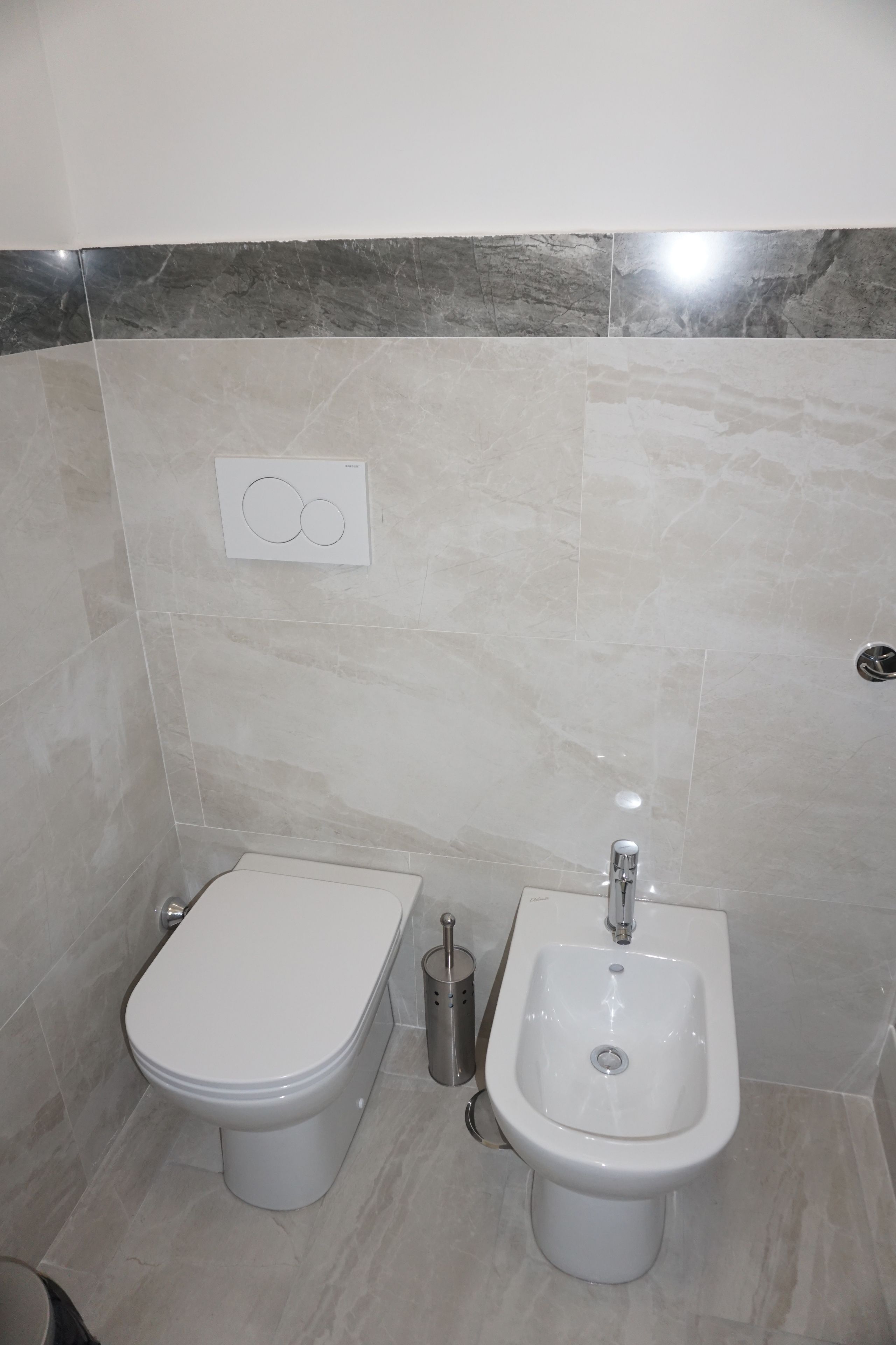 A bathroom with a toilet and bidet, marble tiles, and a wall-mounted flush button.