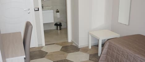Une chambre d’hôtel avec un lit, un bureau, une chaise, une petite table et une salle de bain visible à travers une porte ouverte.