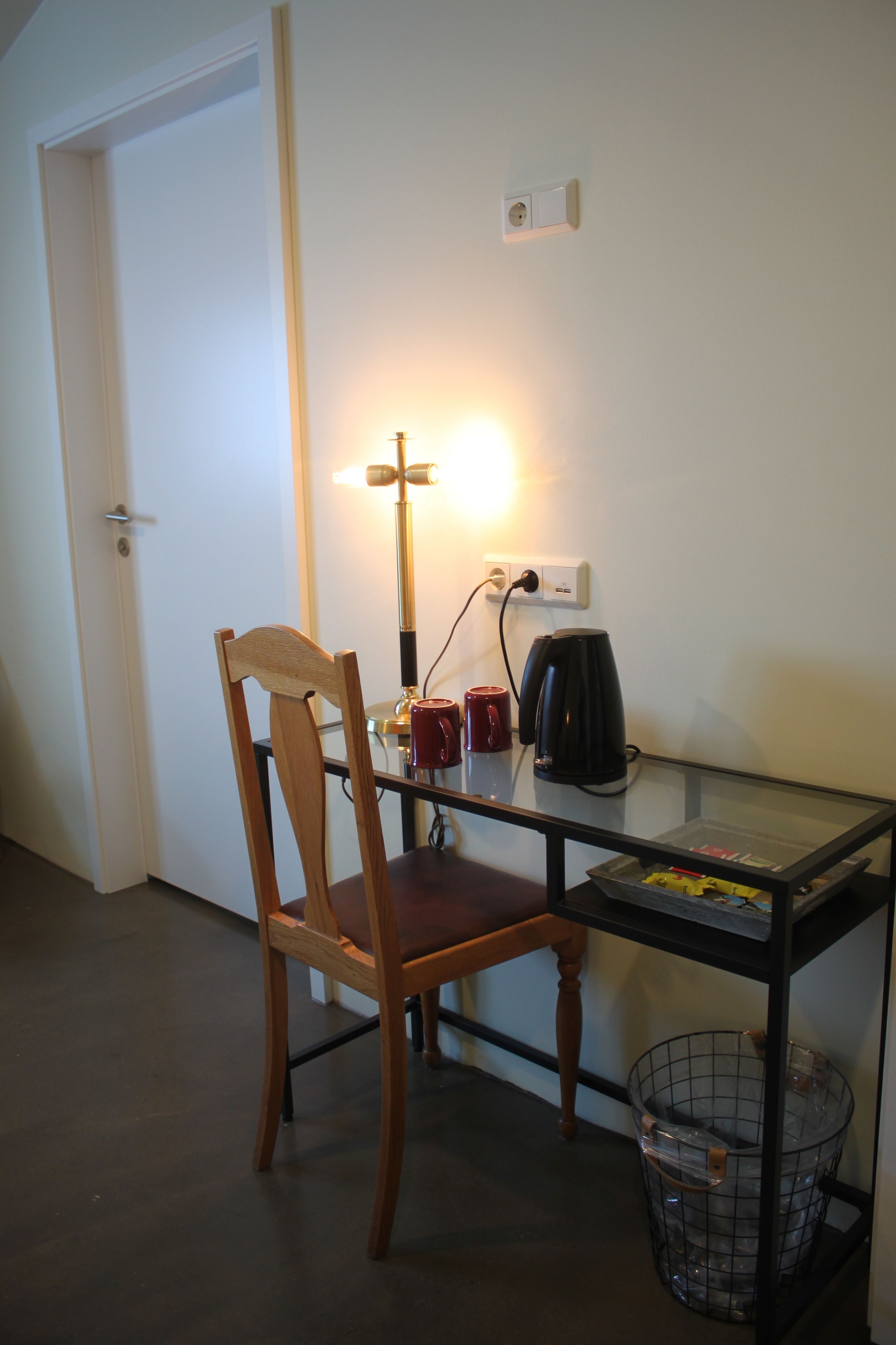 Een kleine, goed verlichte kamer met een houten stoel, een glazen tafel, een zwarte koffiezetapparaat en een prullenbak.