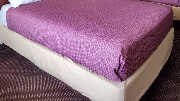 A hotel room with two beds, each with a purple bedspread and white pillows. There are framed artworks on the walls and a nightstand with a lamp between the beds. - Hoosier Inn (Portland)