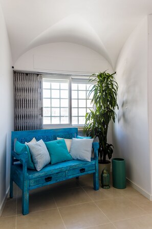 A blue sofa with white and teal cushions, a potted plant, and a green trash bin in a room with white walls and a window with blinds. - Sandhi House - Yoga & Wellness (Mafra)