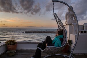 Una persona sentada en una hamaca colgante con vista al mar, cerca hay una estatua de Buda y plantas en macetas.
