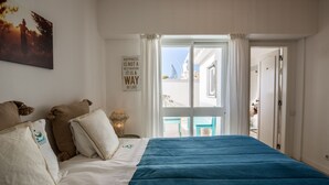 Een slaapkamer met een bed, kussens, een kunstwerk aan de muur, een raam met gordijnen en uitzicht op buiten.