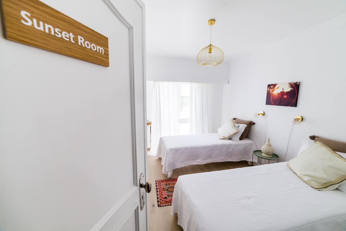Ein Zimmer mit zwei Betten, einem Schild mit der Aufschrift 'Sunset Room', einer Pendelleuchte, einem Wandbild und einem kleinen Tisch mit einer Lampe.