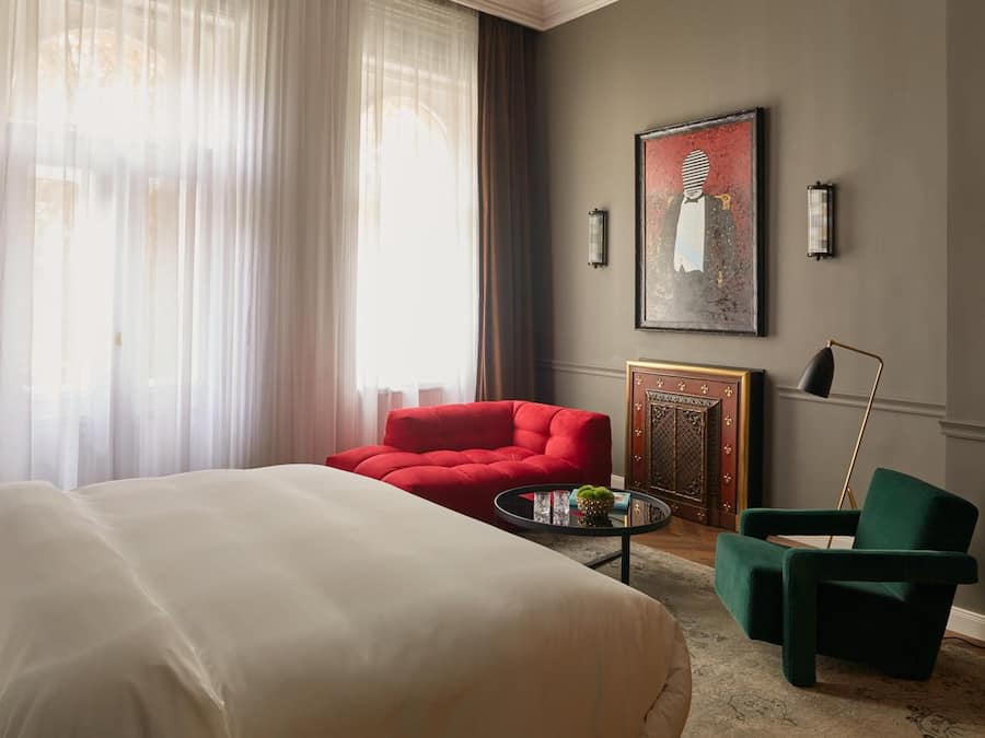 Ein Hotelzimmer mit einem großen Bett, einem roten Sessel, einem grünen Sessel, einem kleinen runden Tisch mit einer Pflanze und einem gerahmten Bild an der Wand.