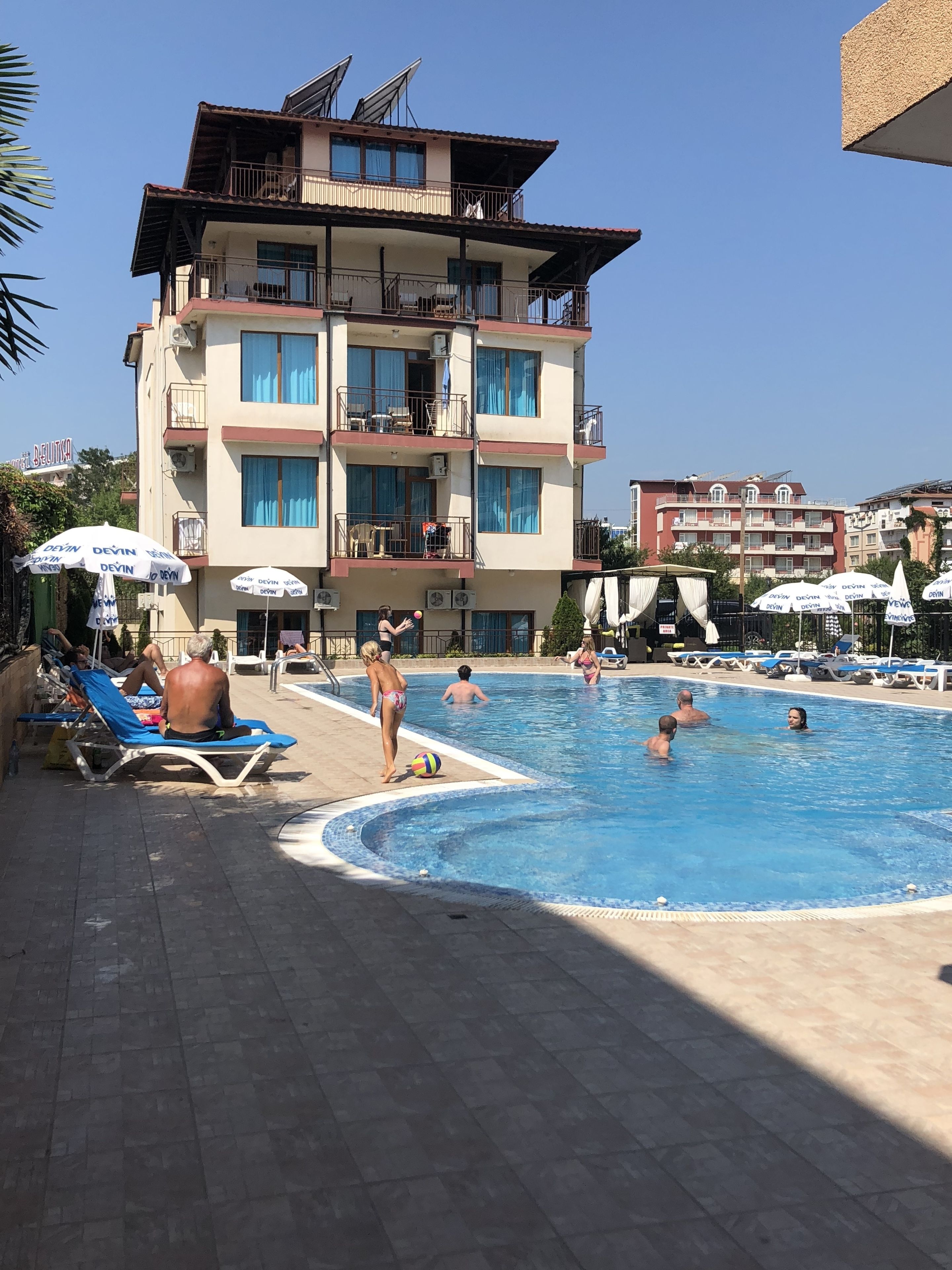 A swimming pool area with lounge chairs and umbrellas, a multi-story building with balconies, and a clear sky.