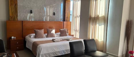 Una habitación de hotel con una cama grande, un cabecero de madera, una mesita de noche con un teléfono, un escritorio con una silla y una mesa de centro de vidrio.