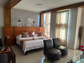 Una habitación de hotel con una cama grande, un cabecero de madera, una mesita de noche con un teléfono, un escritorio con una silla y una mesa de centro de vidrio.