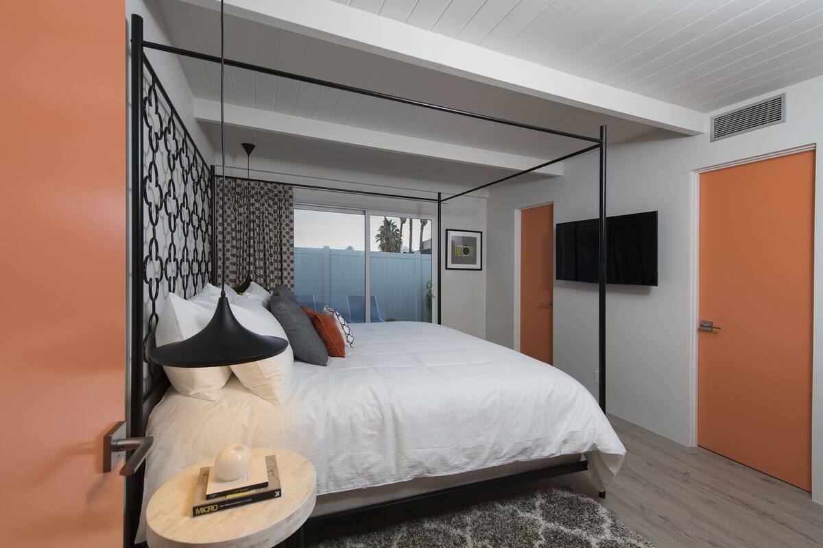 A modern bedroom with a large bed, a bedside table with a lamp, a TV mounted on the wall, and a view of the outdoors.