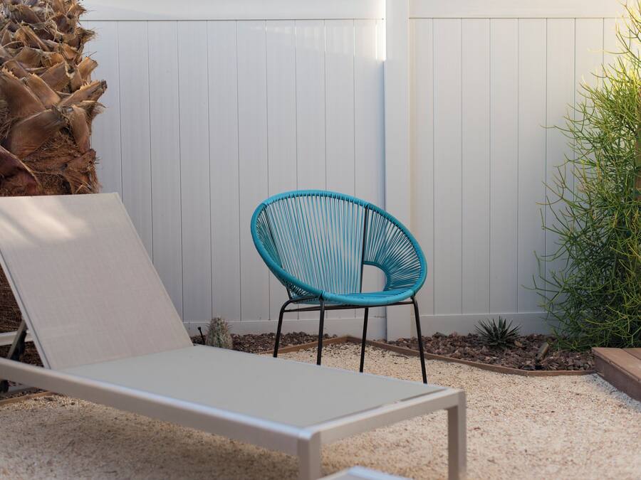 A white lounge chair, a blue woven chair, a palm tree, and a white fence in a garden setting.