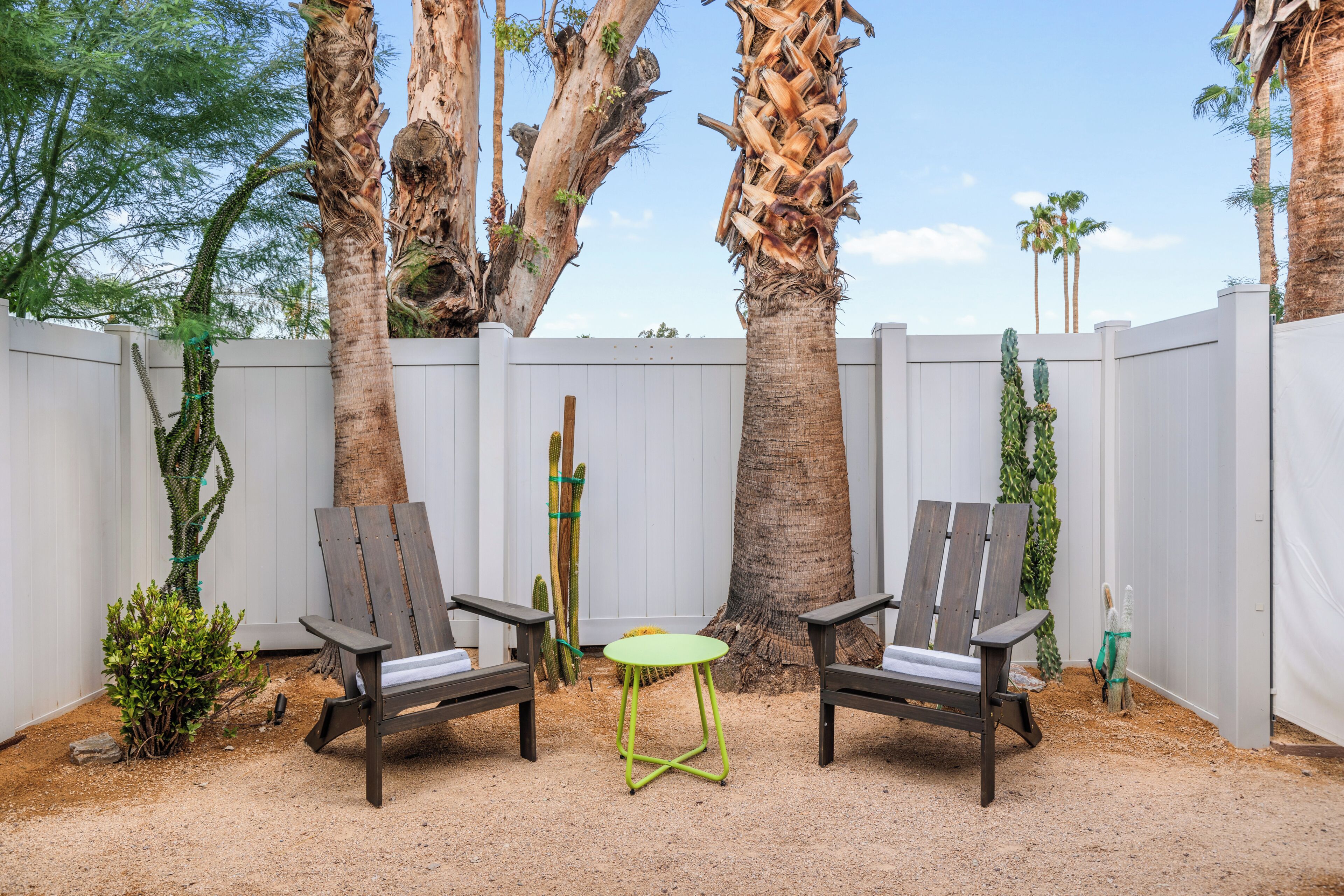 Une terrasse avec deux chaises en bois et une petite table verte, entourée d’une clôture blanche et de palmiers.