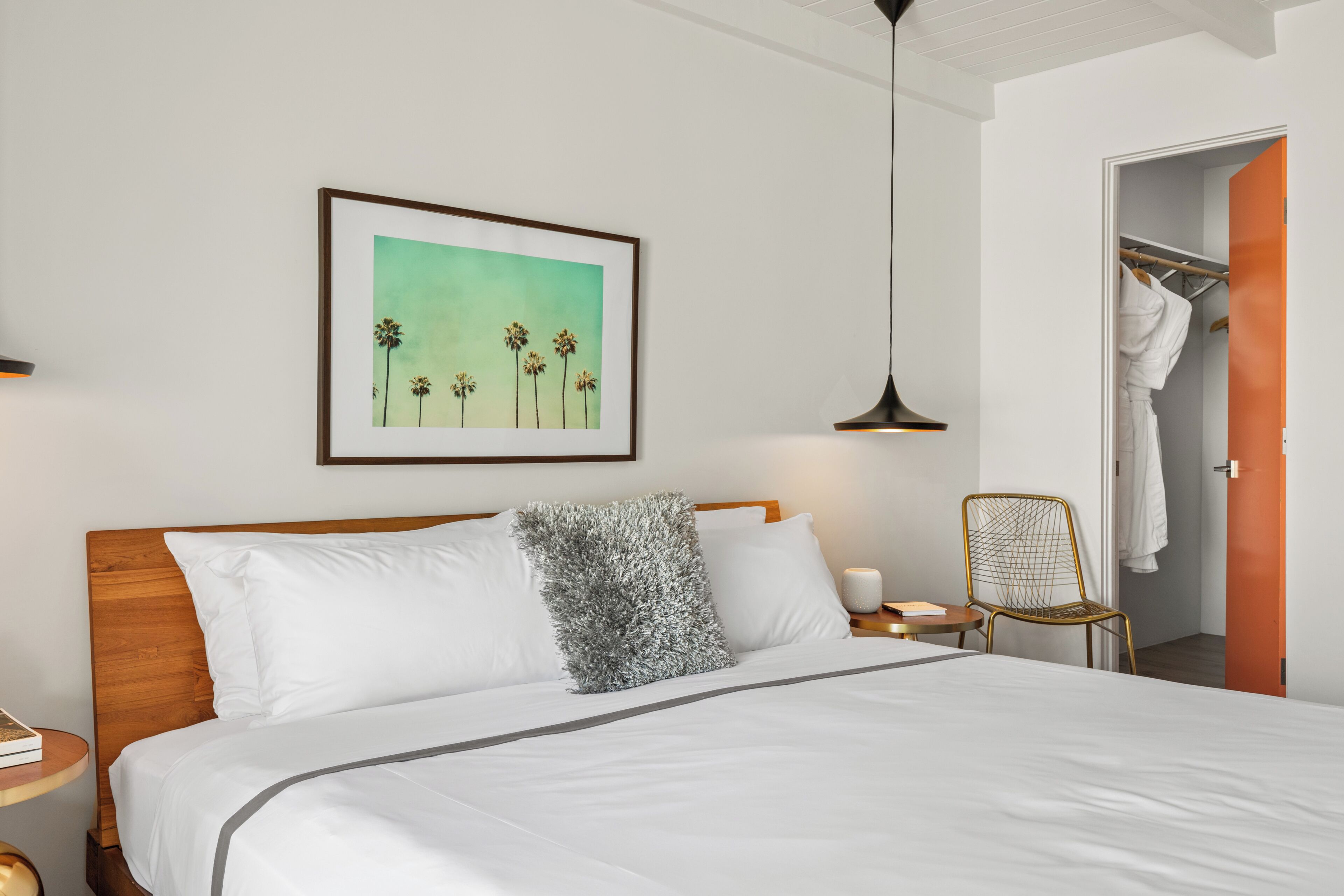 A neatly made bed with white linens, a framed picture of palm trees, a hanging lamp, and a chair.