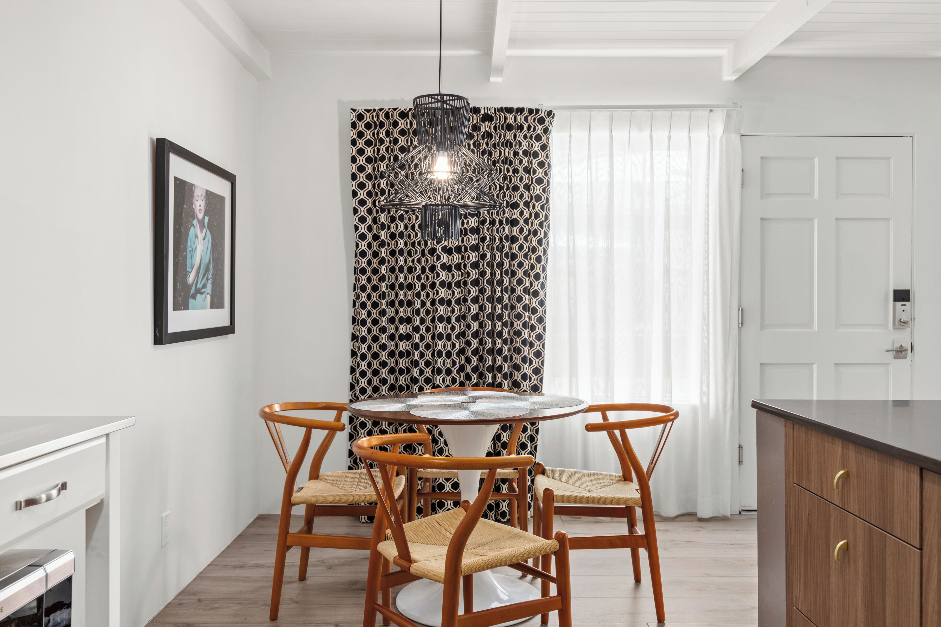 Un coin repas avec une table ronde, quatre chaises en bois, un rideau à motifs, un luminaire suspendu et une porte blanche.