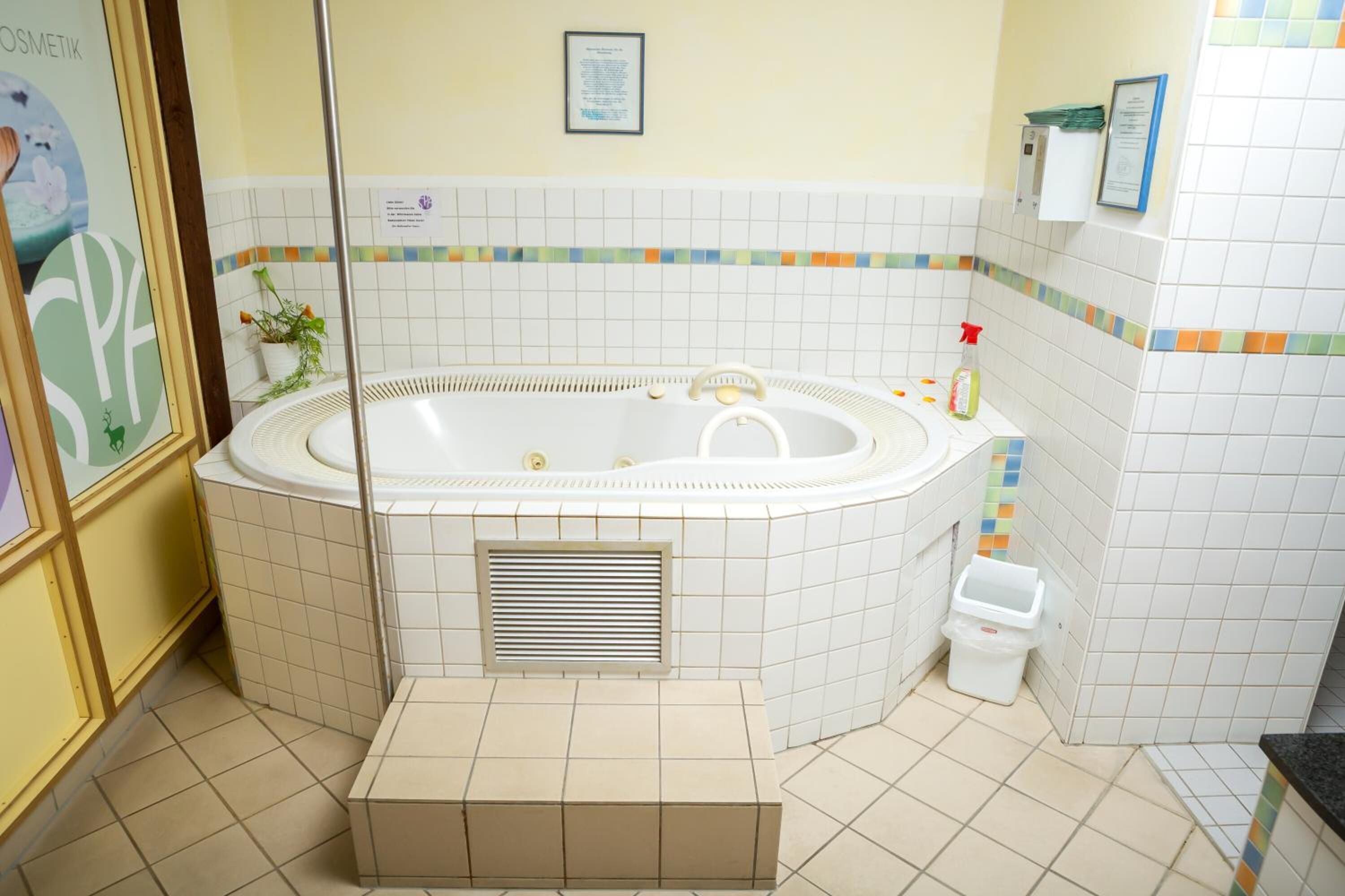 Ein Badezimmer mit einer großen Badewanne, einer Dusche, einem Waschbecken und einer Toilette. Ein kleiner Mülleimer und ein gerahmtes Zertifikat befinden sich an der Wand.