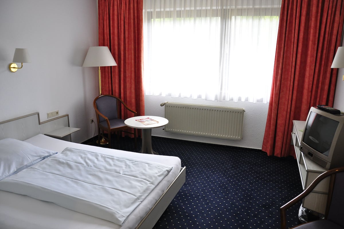 Une chambre d’hôtel avec un lit, une chaise, une petite table, un téléviseur et une fenêtre avec des rideaux.