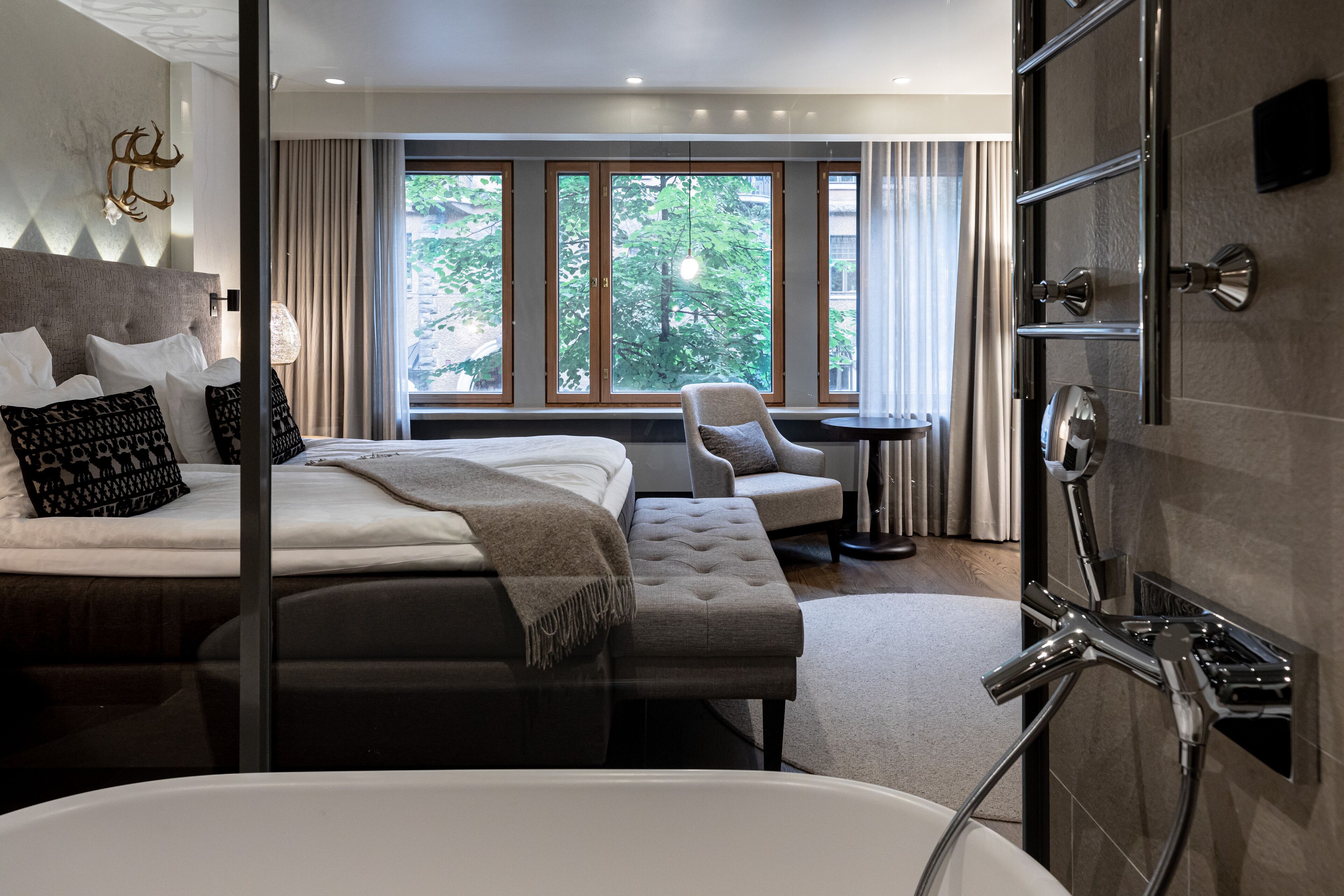 Quarto de hotel moderno com uma cama grande, área de estar, banheira e vista para o verde através de amplas janelas.