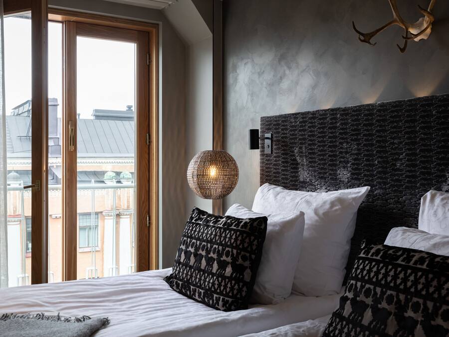A modern hotel room with a large bed, patterned pillows, a textured wall, and a view through a sliding door.