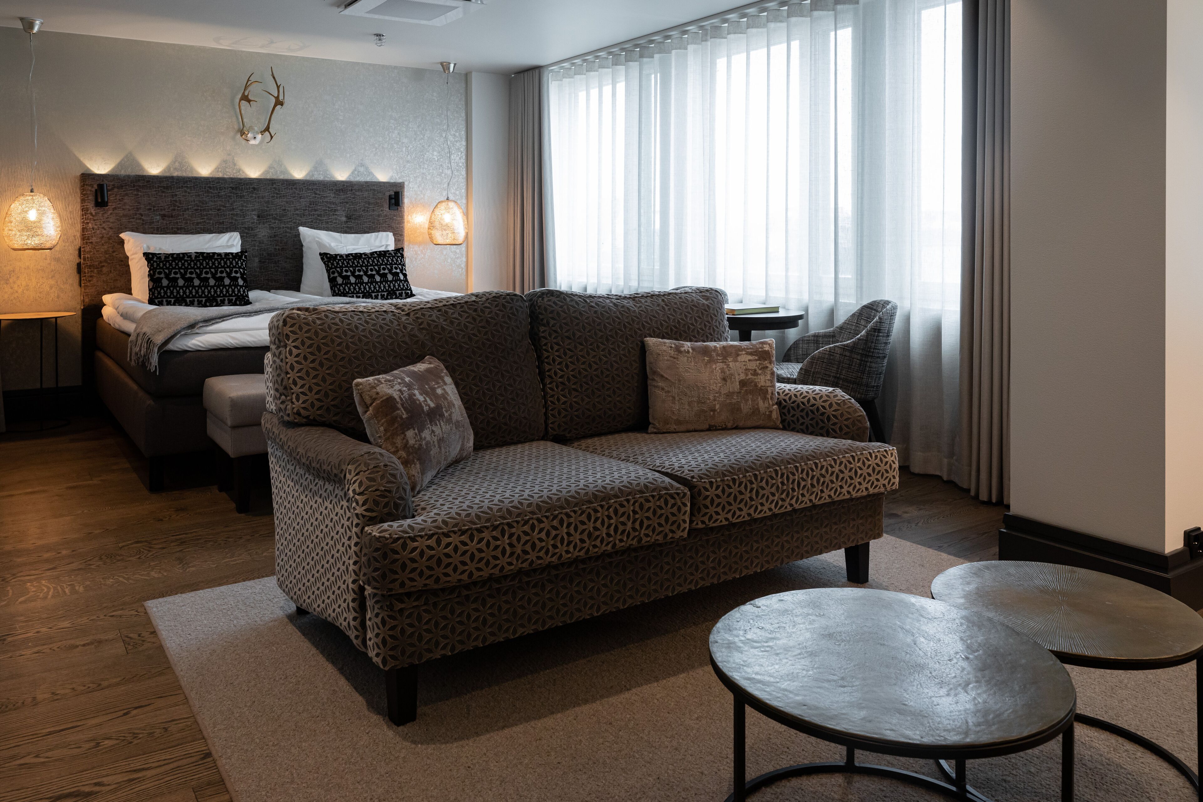 A modern hotel room with a patterned sofa, a bed with patterned pillows, a small table, and a decorative wall piece.