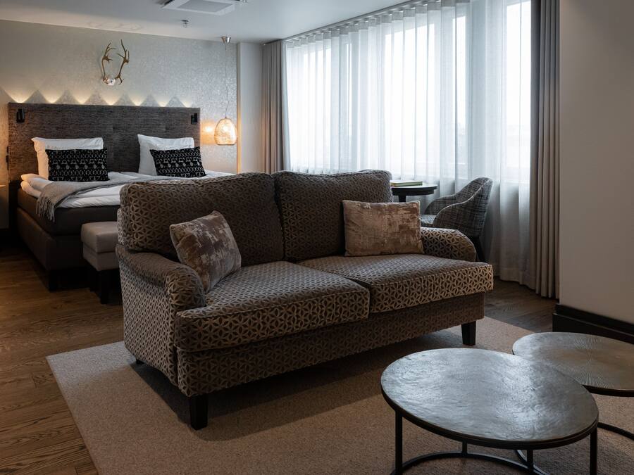 A modern hotel room with a patterned sofa, a bed with patterned pillows, a small table, and a decorative wall piece.
