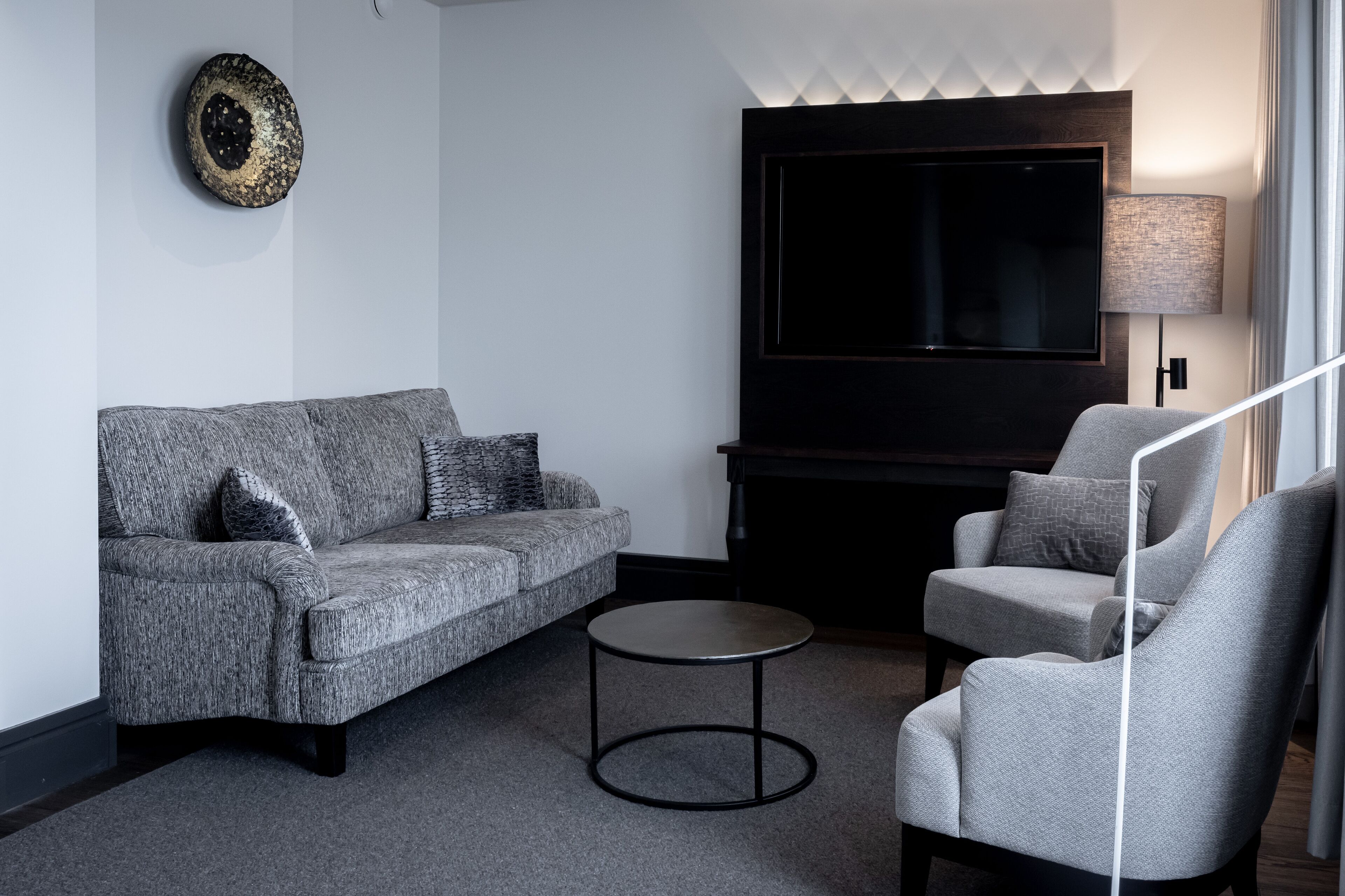 Un salon moderne avec un canapé gris, un fauteuil gris, une table basse ronde, un téléviseur à écran plat fixé au-dessus d’une cheminée et une lampe murale.