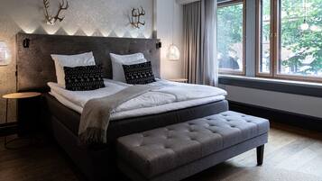 A modern bedroom with a large bed, a grey upholstered bench, a headboard with wall-mounted decor, and a window with a view of greenery.