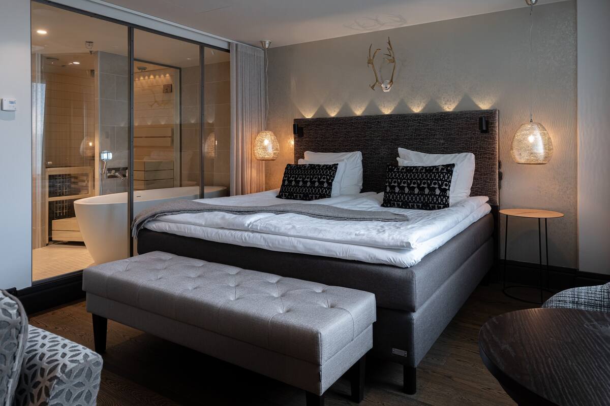 A modern bedroom with a large bed, a bench, a small table, and a glass shower area.