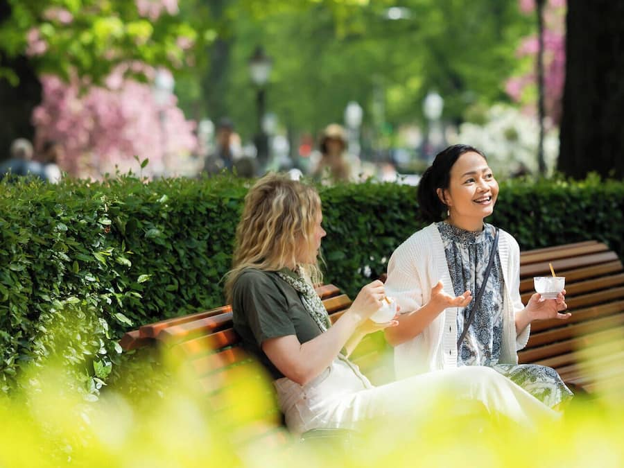 Two people sitting on a bench in a park with greenery and trees in the background.